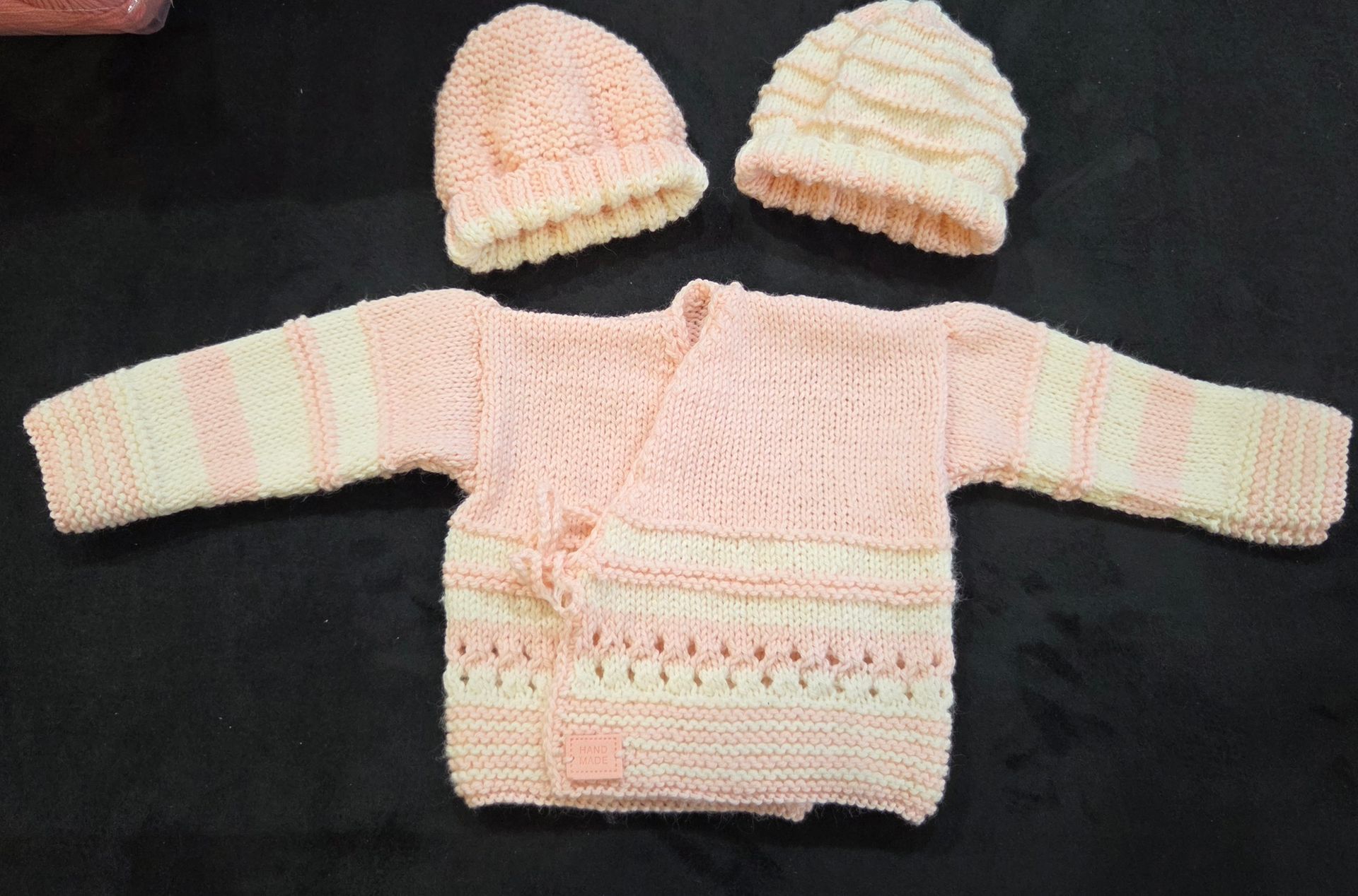 Gilet bébé tricoté à la main, couleur crème et rose clair, avec deux bonnets assortis sur fond foncé.
