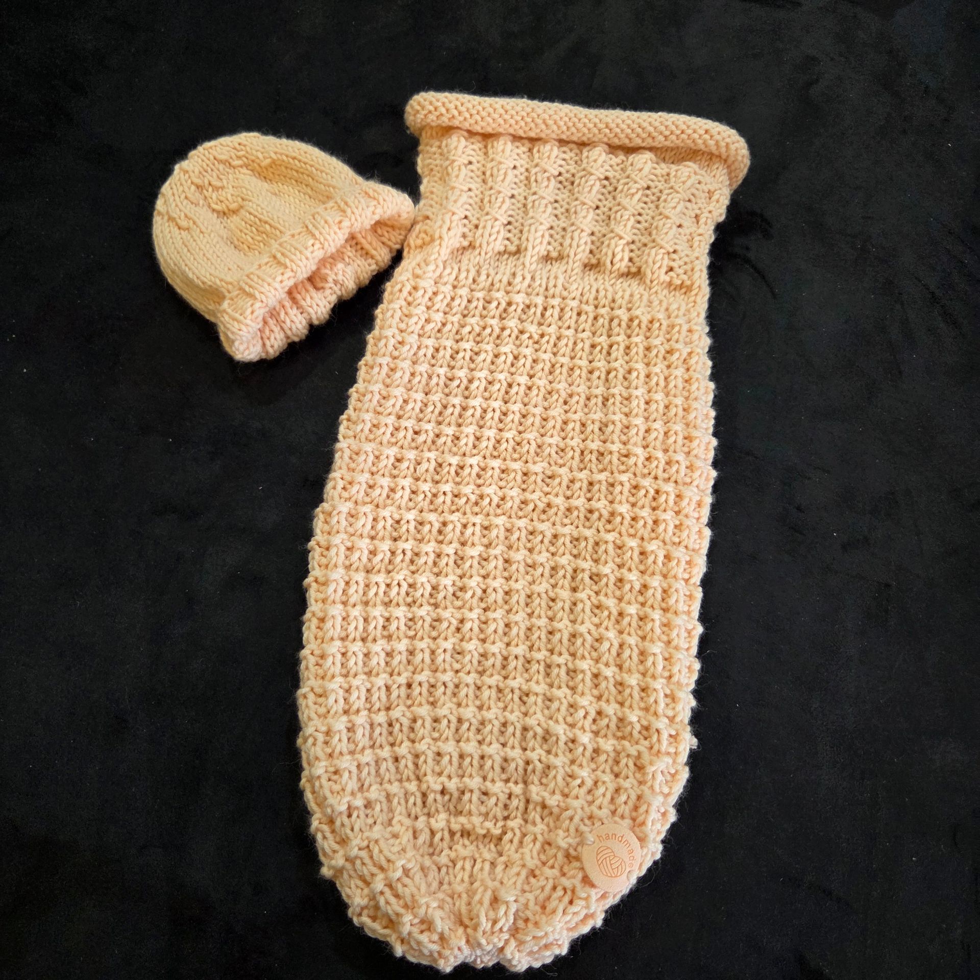 Un sac de couchage cocon tricoté couleur pêche pour bébé et un petit bonnet assorti, disposés sur un fond sombre.