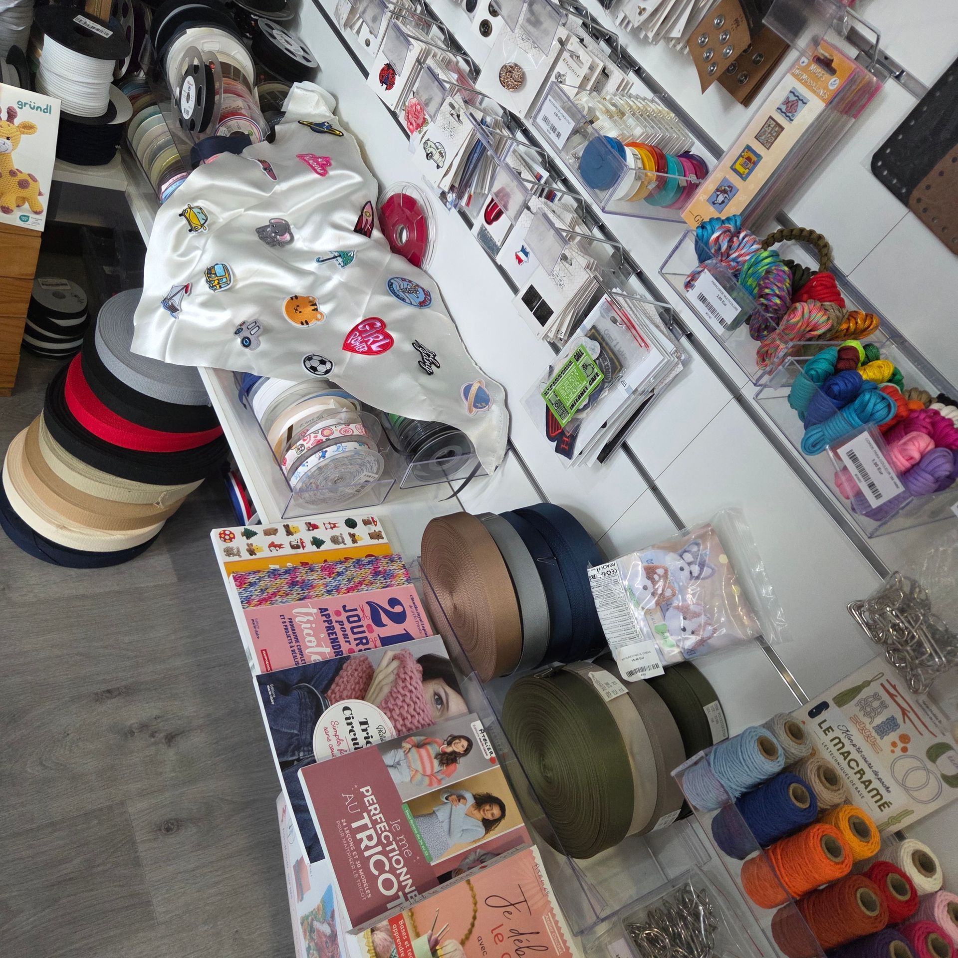 Une vitrine de magasin de loisirs créatifs présentant des rubans, des tissus à motifs, des livres de loisirs créatifs et des bobines de fil sur des étagères blanches.