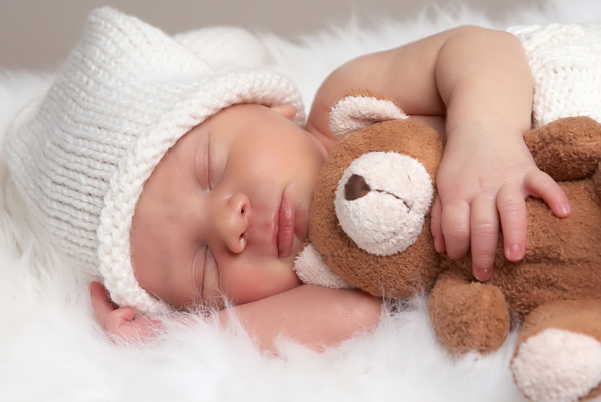 Un bébé endormi porte un bonnet blanc en tricot et repose sur une couverture en fausse fourrure blanche, serrant contre lui un petit ours en peluche brun.