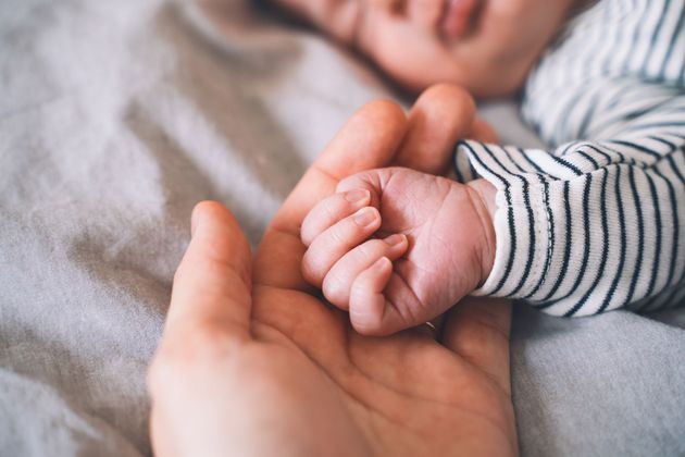 La main d'un tout petit bébé repose dans la paume d'un adulte, sur un fond de tissu gris clair.