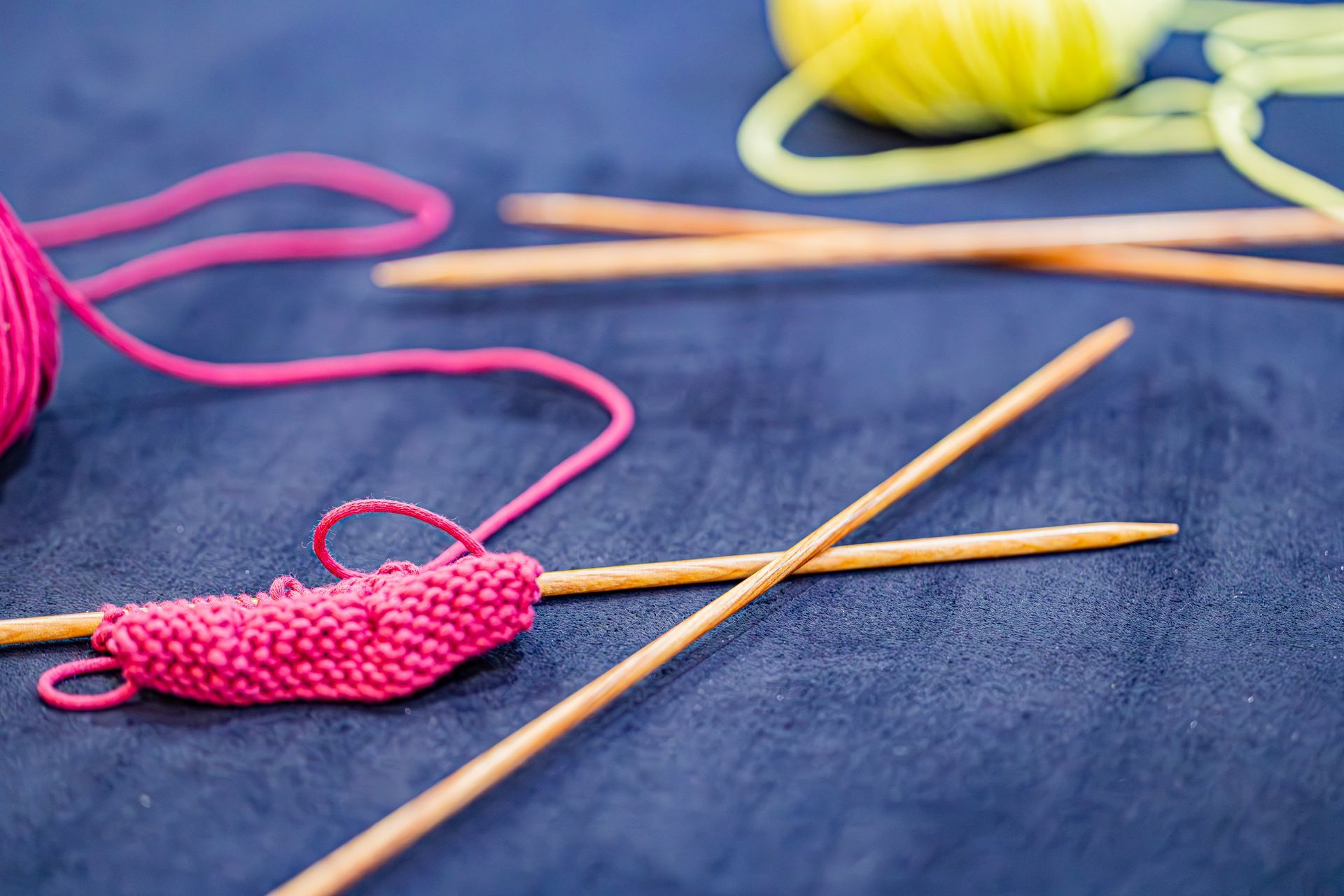 Tricot rose vif sur aiguilles en bambou avec une pelote de laine jaune en arrière-plan sur fond bleu.