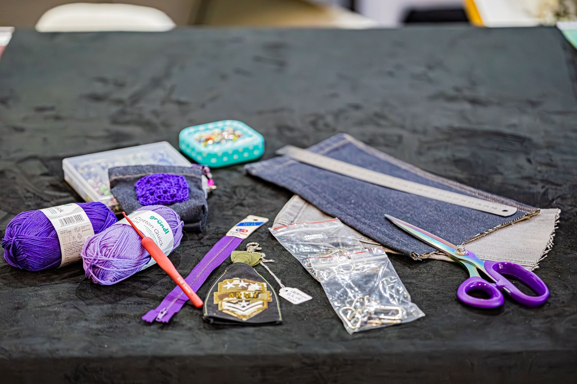 Matériel de loisirs créatifs, notamment du fil violet, un crochet, du tissu denim, des ciseaux et des articles de couture, sur une surface noire.