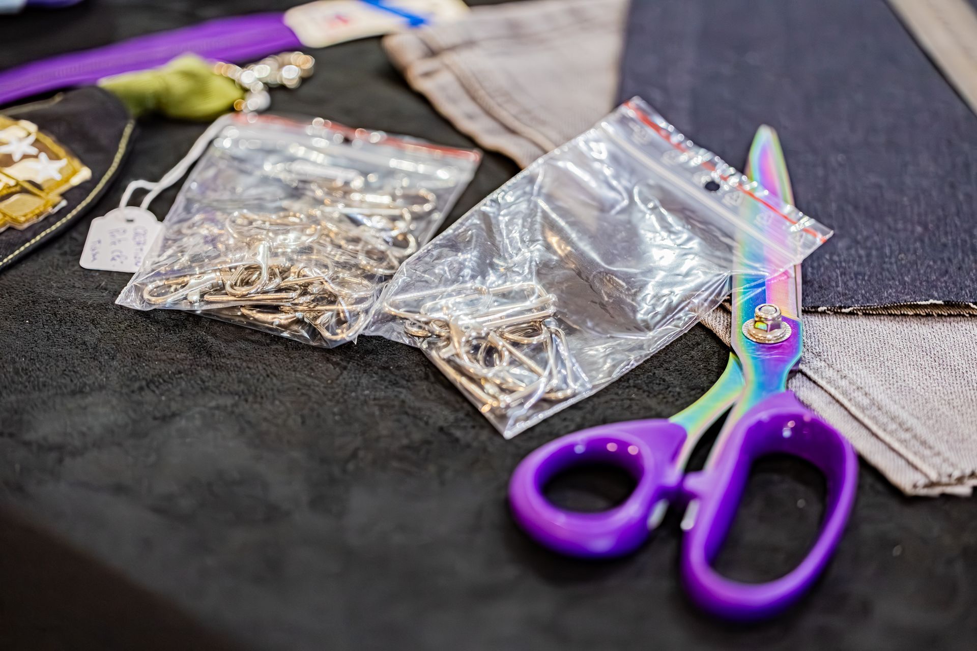 Des ciseaux aux couleurs de l'arc-en-ciel et aux poignées violettes sont posés sur une table à côté d'un sac en plastique contenant des chaînes métalliques et des chutes de tissu.