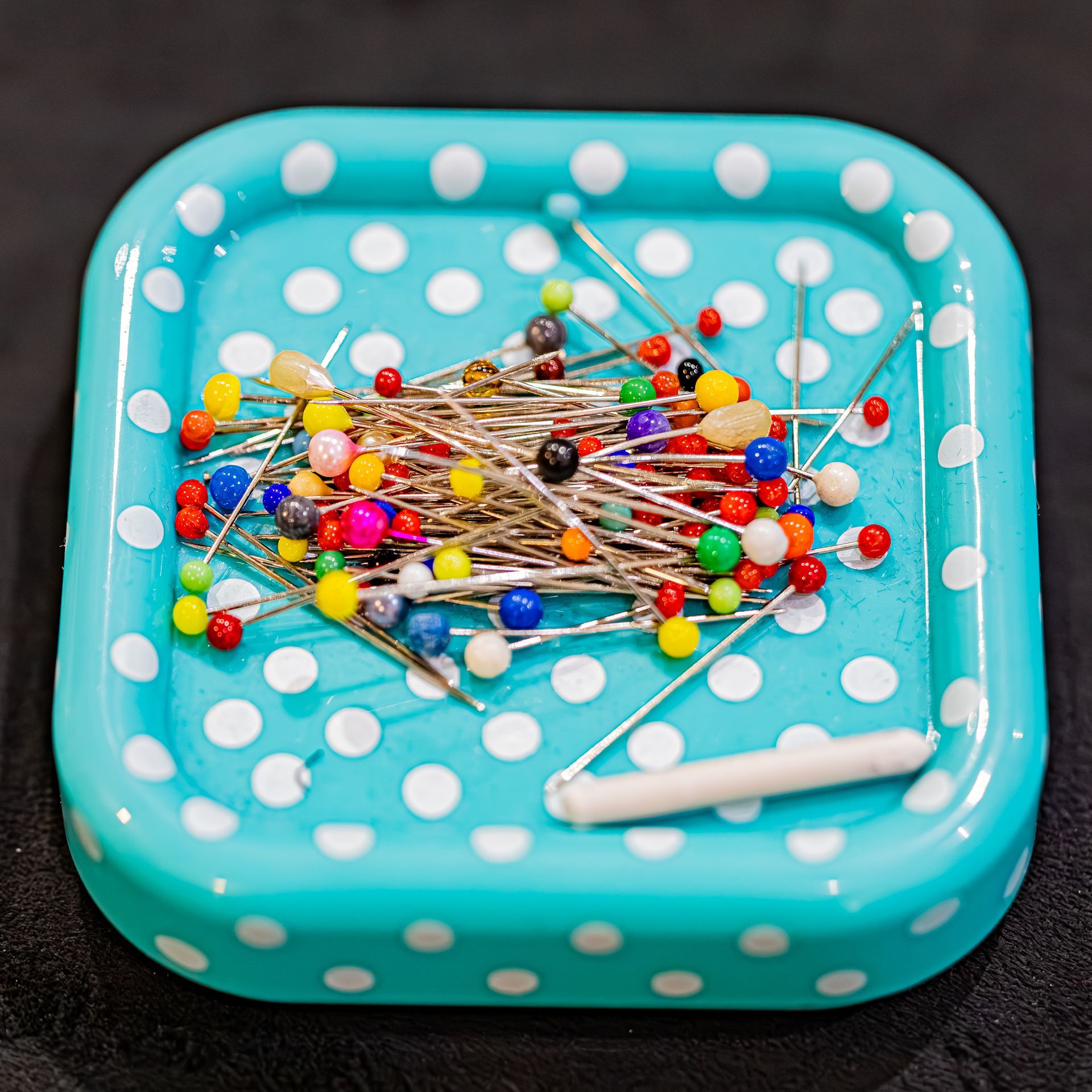 Un petit plateau carré turquoise à pois blancs, rempli d'un assortiment d'épingles à coudre multicolores.
