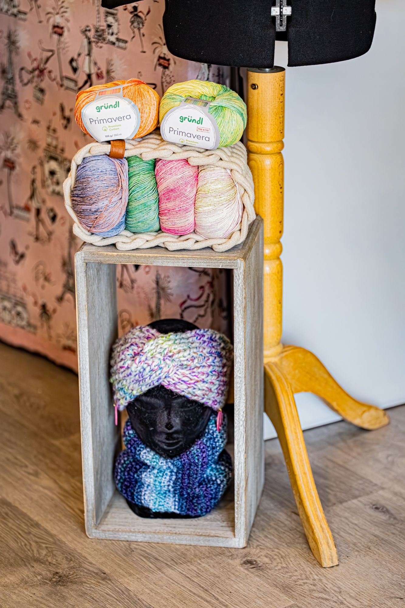 Une caisse en bois contenant des écheveaux de laine est posée sur un support de mannequin, la tête de ce mannequin portant un bandeau et un col tricotés.