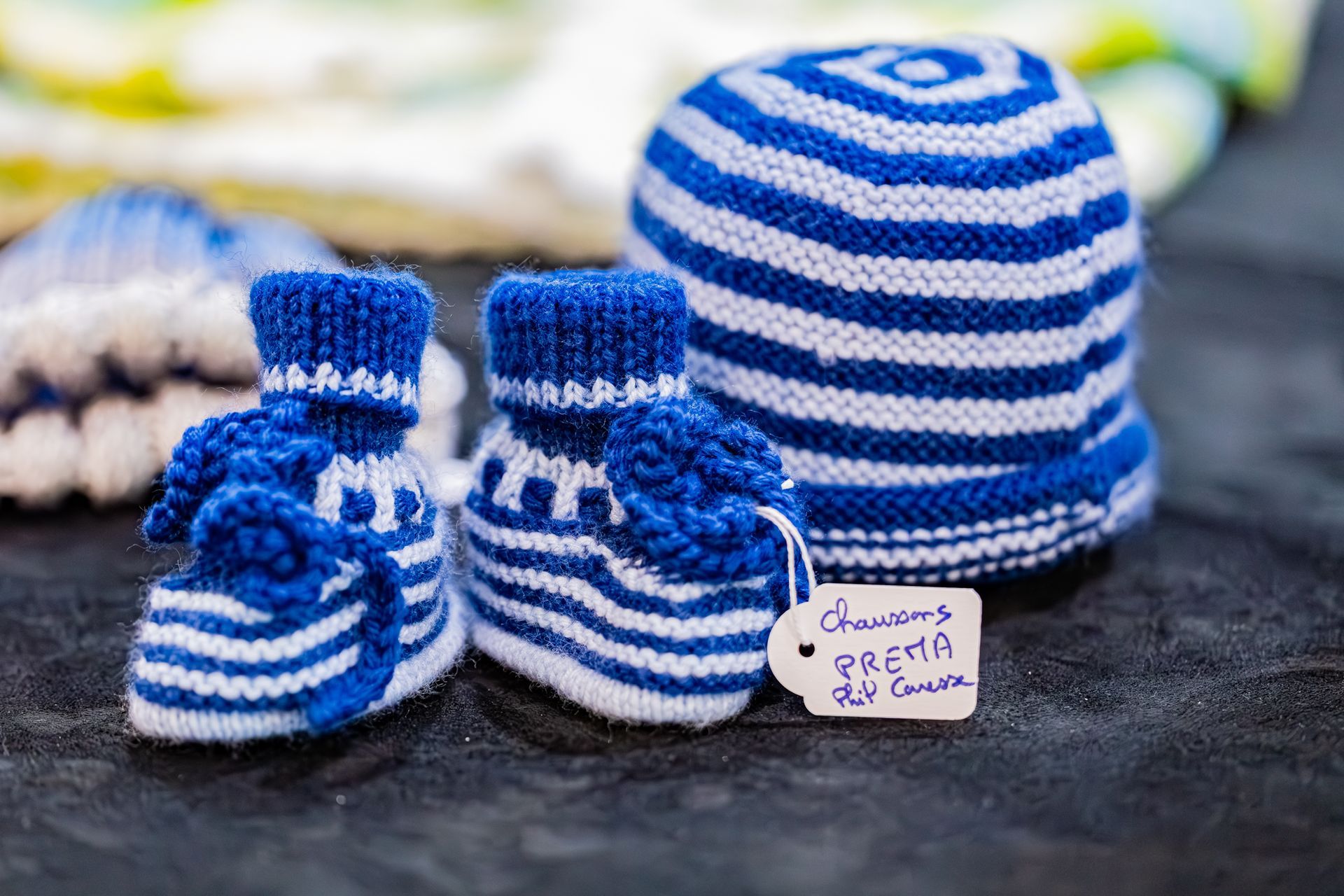 Des chaussons de bébé en tricot à rayures bleues et blanches et un bonnet assorti sont posés sur une surface sombre, une étiquette étant attachée à l'un des chaussons.