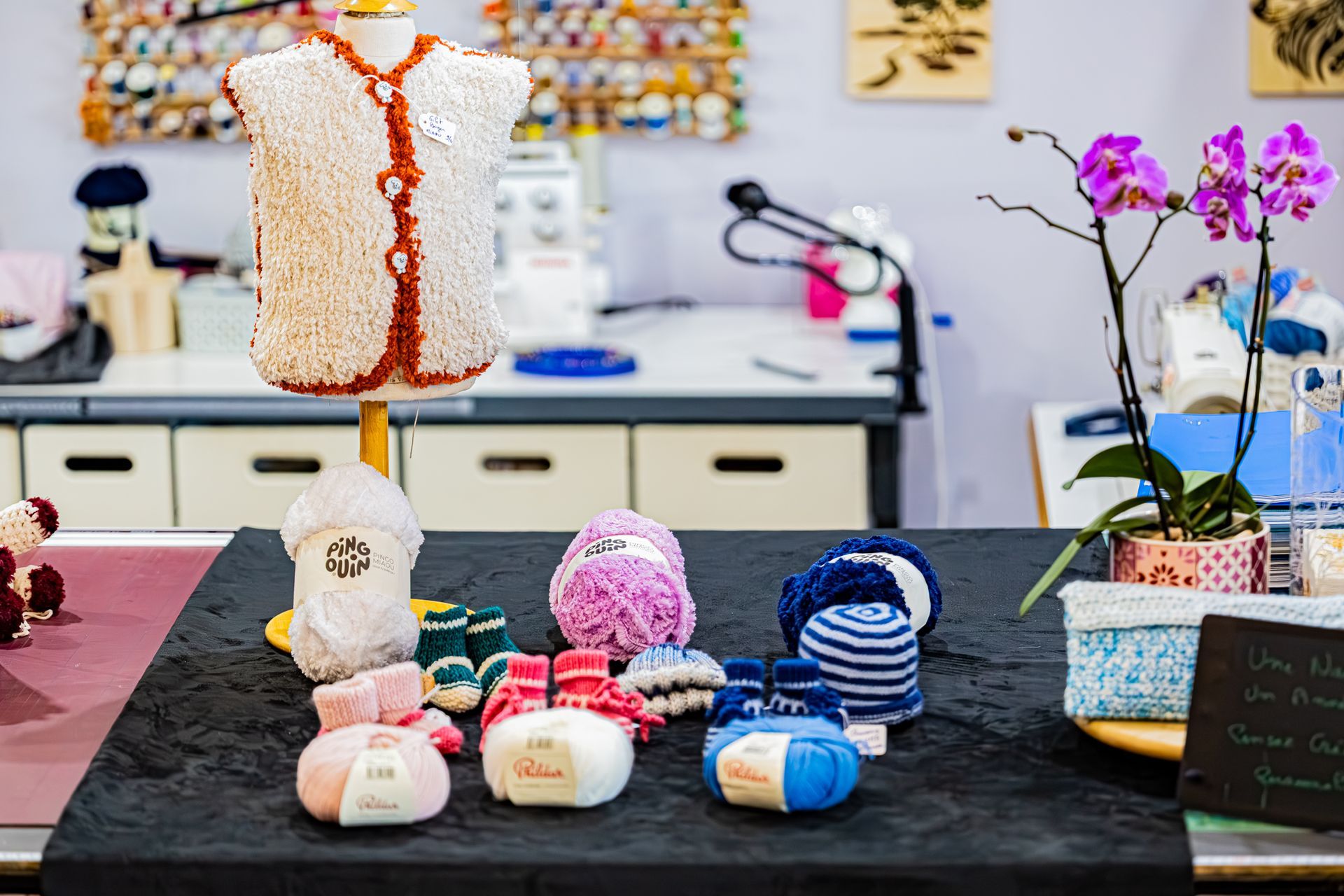 Sur une table d'atelier de loisirs créatifs, on trouve un mannequin portant un gilet tricoté, diverses pelotes de laine colorée, des chaussons de bébé et des orchidées.