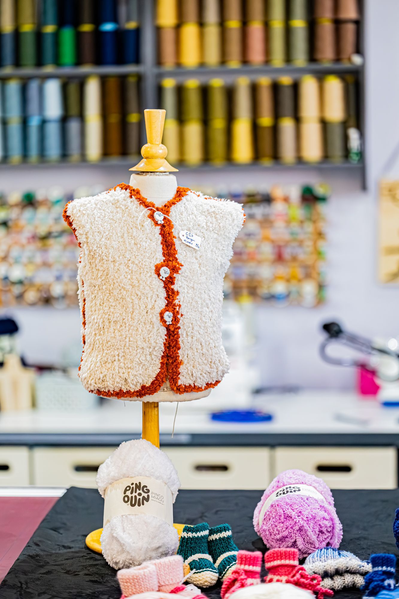 Un mannequin vêtu d'un gilet crème duveteux à bordures orange se tient sur une table avec des pelotes de laine et de petits chaussons de bébé tricotés.