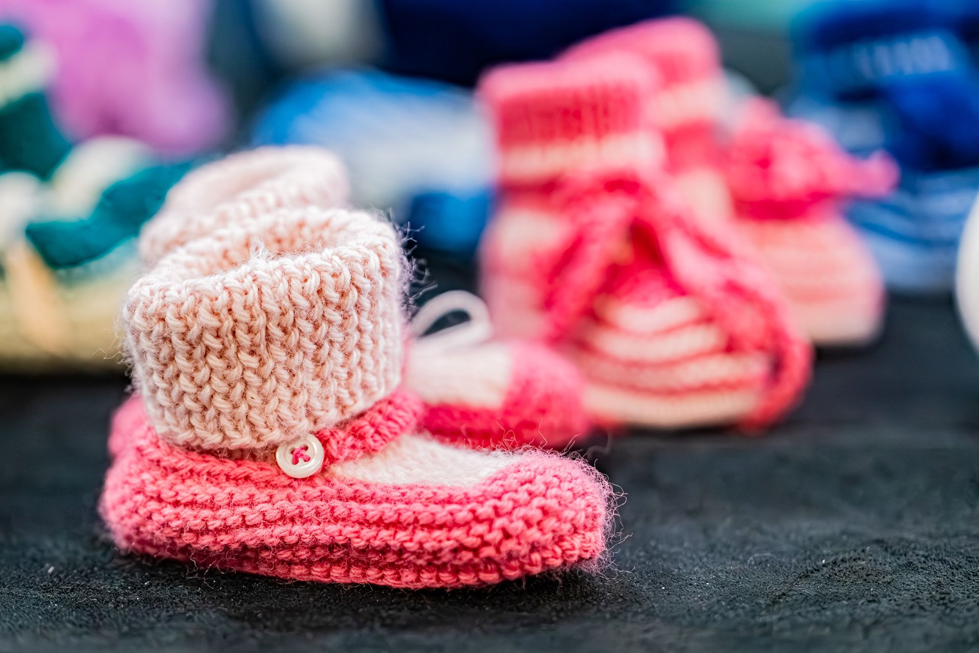 Gros plan sur une paire de petits chaussons de bébé roses et beiges tricotés à la main, avec de minuscules boutons blancs sur une surface foncée.