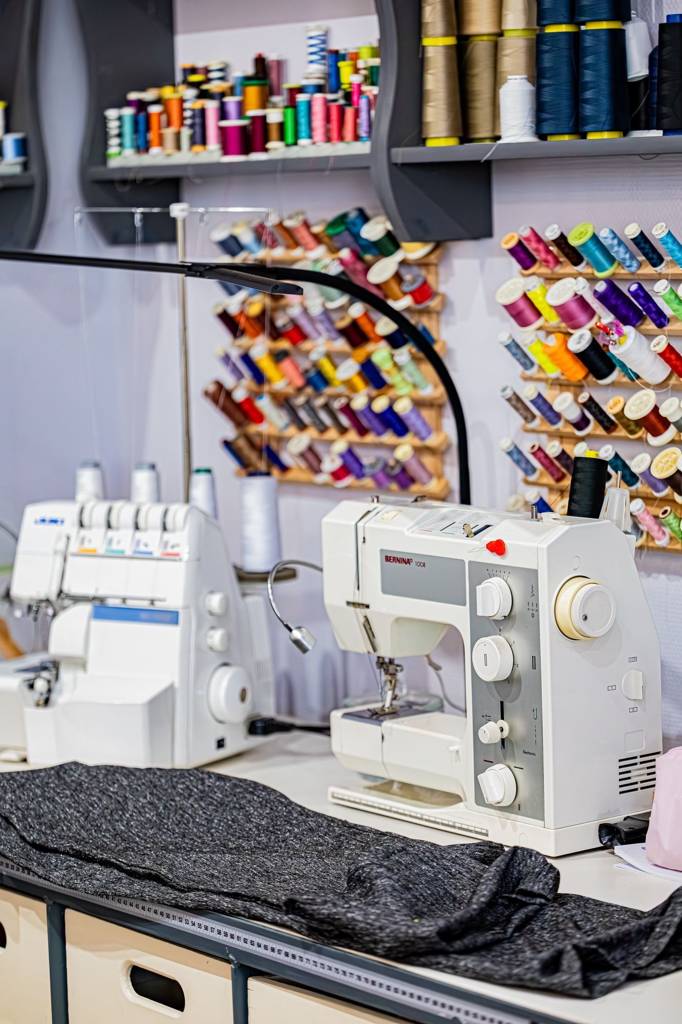 Un poste de couture comprenant deux machines, un morceau de tissu gris et des murs recouverts d'étagères et de supports à bobines de fil.