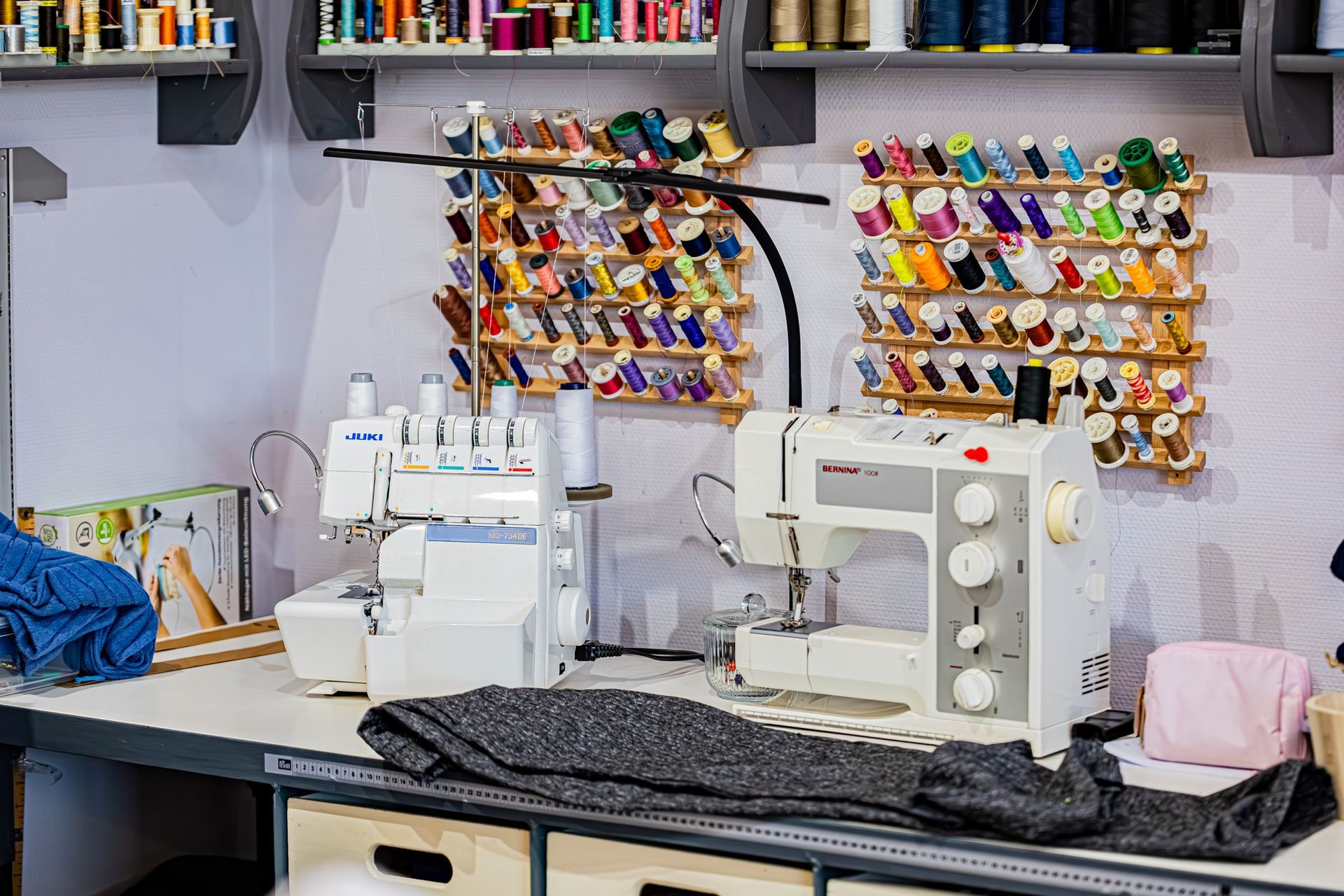 Un espace de couture comprend une surjeteuse et une machine à coudre sur une table, avec des bobines de fil exposées sur des étagères murales au-dessus.