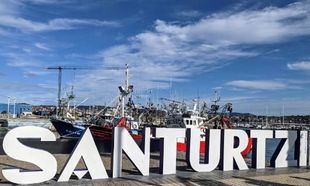 Grandes letras blancas SANTURTZI frente a barcos en un puerto bajo un cielo azul.