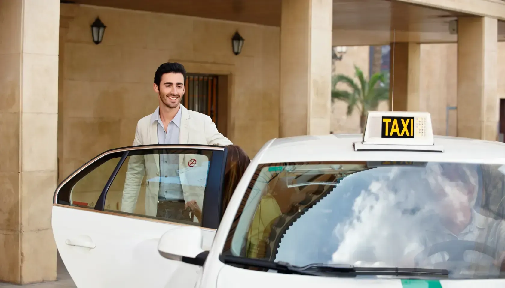 Un hombre sonríe junto a un taxi blanco descapotable frente a la entrada de un edificio.