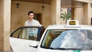 Un hombre sonríe junto a un taxi blanco descapotable frente a la entrada de un edificio.