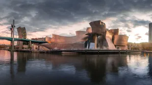 Edificio moderno frente al mar con torres curvas de color tostado que se reflejan en las tranquilas aguas al atardecer.