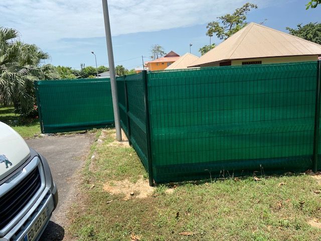 Clôture en grillage vert le long d'une allée, avec une voiture au premier plan et des bâtiments à l'arrière-plan.