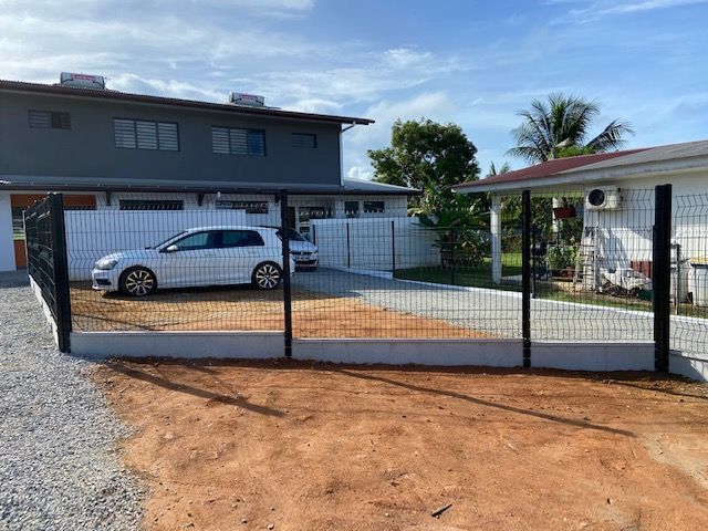 Une clôture en grillage noir entoure une allée où une voiture blanche est garée sur du gravier.