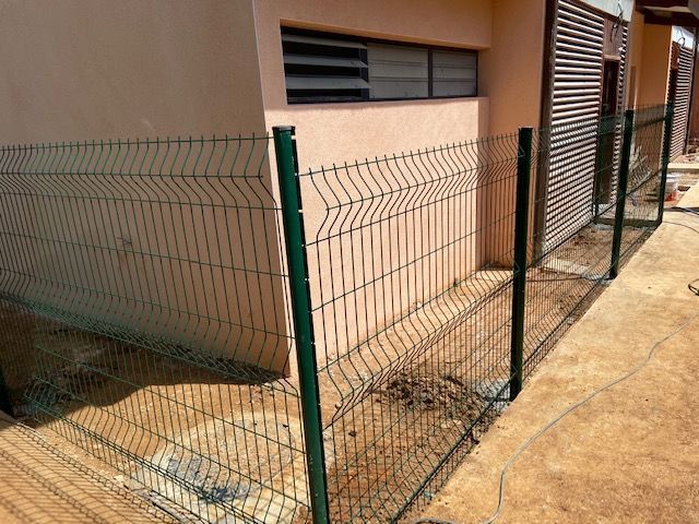 Clôture en grillage vert longeant le mur beige d'un bâtiment et le sol en béton.