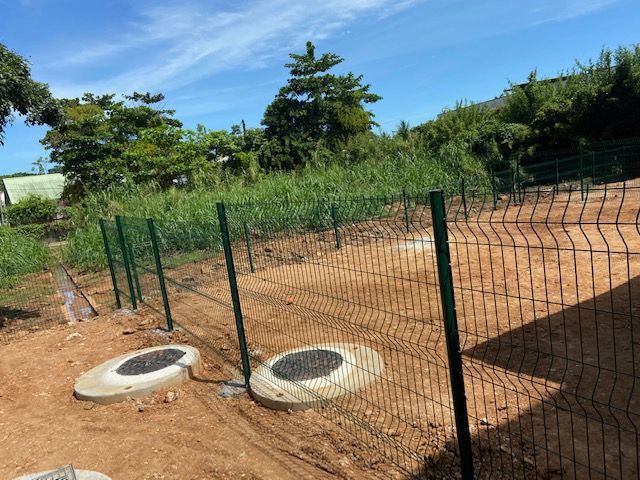 Clôture en fil de fer vert, plaques d'égout en béton et chemin de terre dans un cadre extérieur ensoleillé.