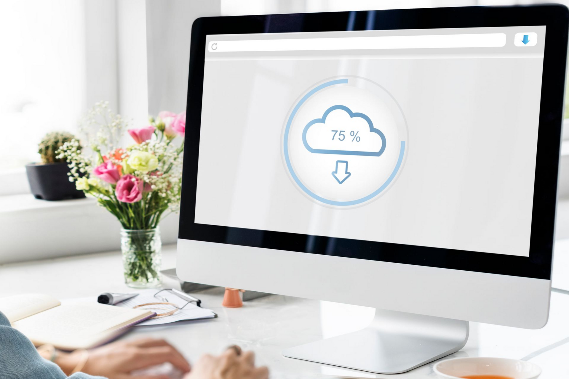 Écran d'ordinateur affichant une icône de téléchargement cloud sur un bureau lumineux, à côté de fleurs et d'une tasse de café.
