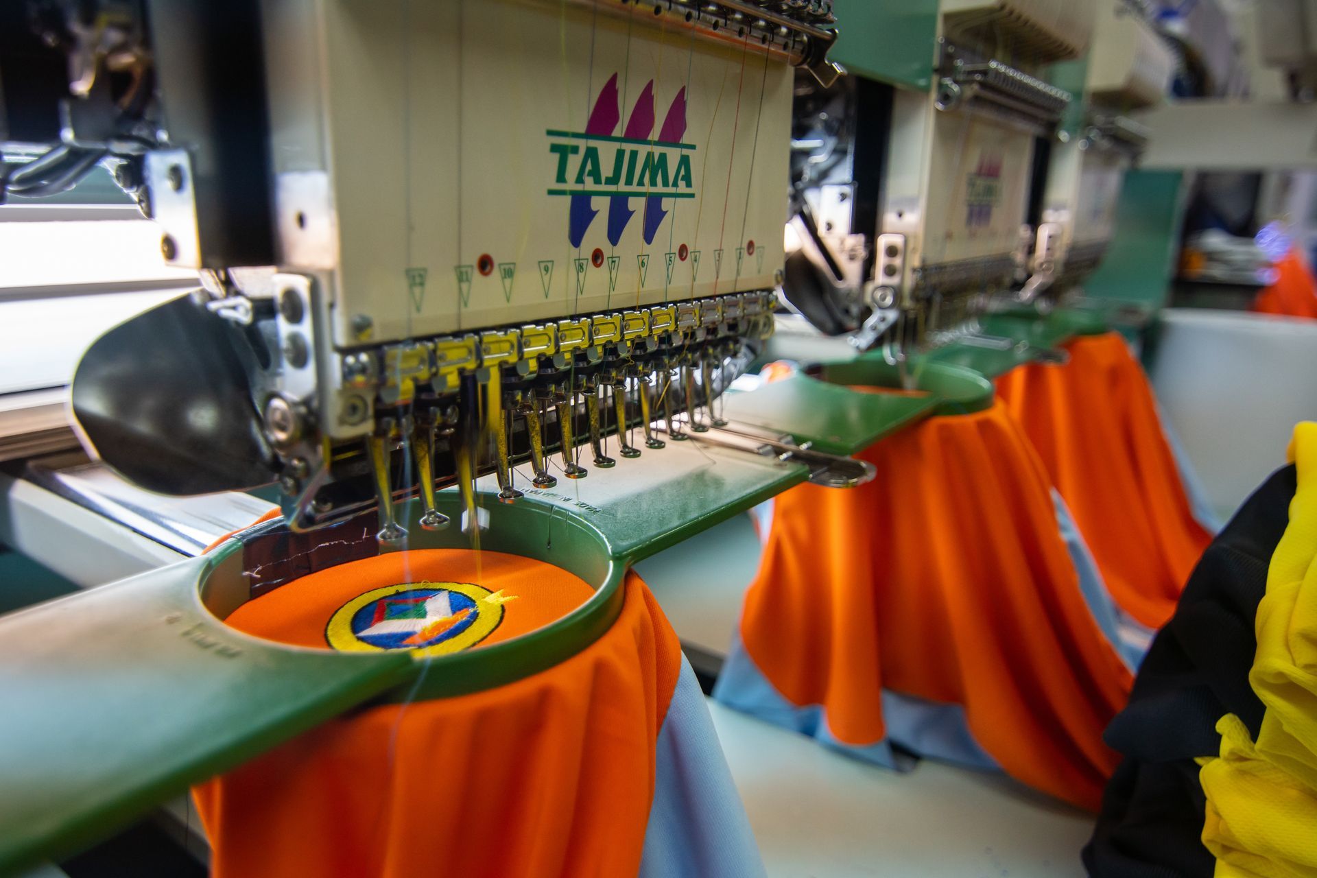 Machine à broder cousant un tissu orange sur un panneau de vêtement circulaire avec un logo multicolore