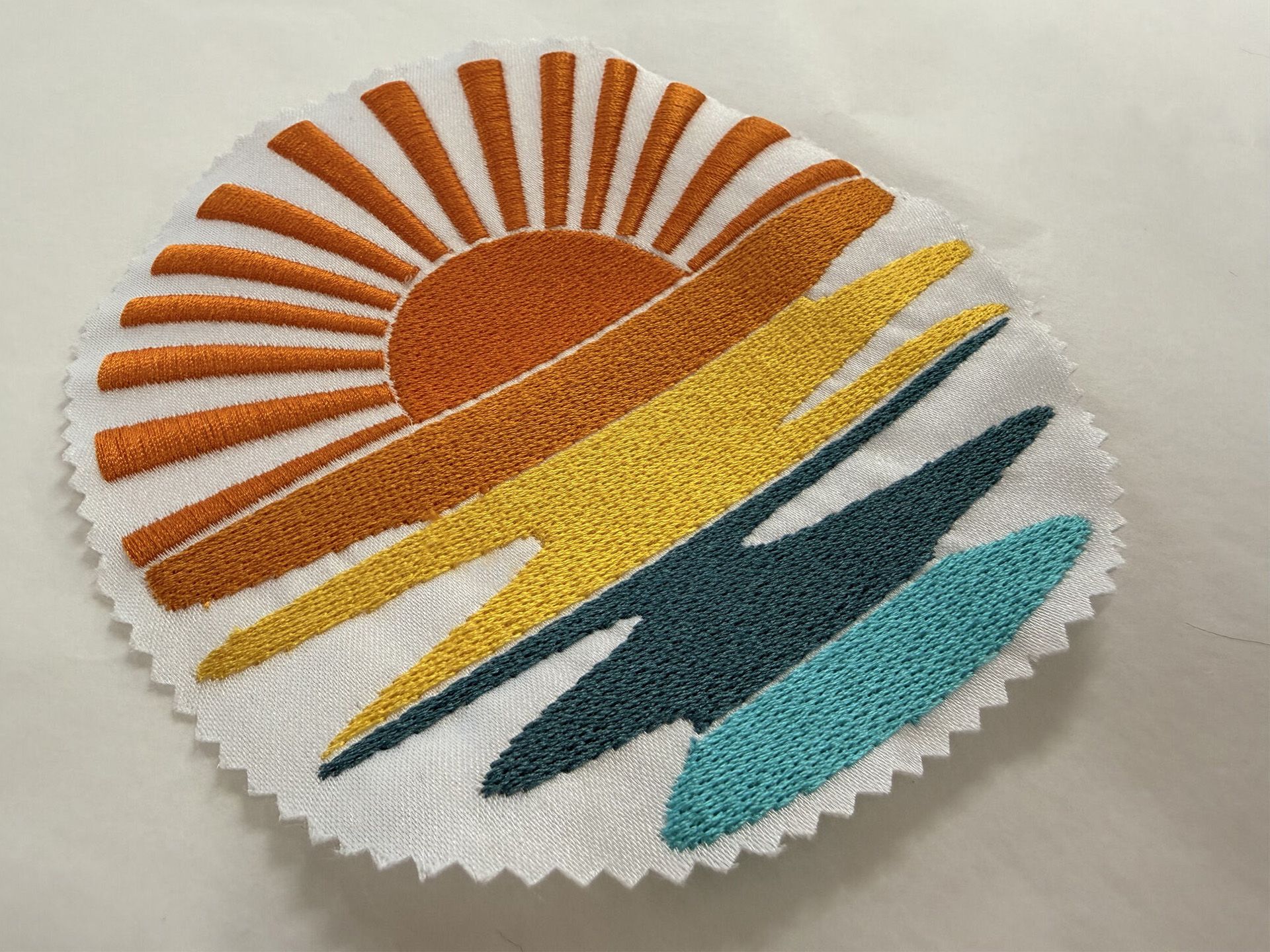 Écusson rond brodé représentant un lever de soleil orange, jaune et turquoise sur tissu blanc
