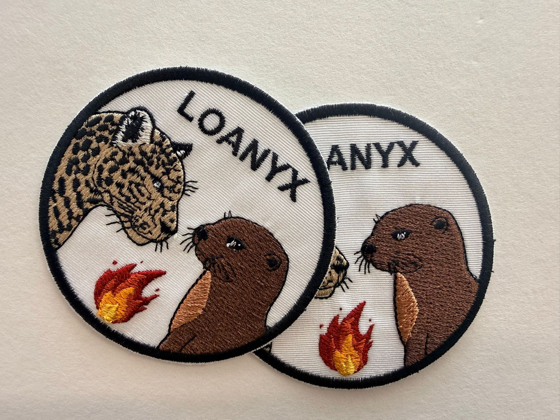 Deux écussons ronds brodés avec des têtes d'animaux, une icône de flamme et les inscriptions « LOANYX » et « ANYX ».