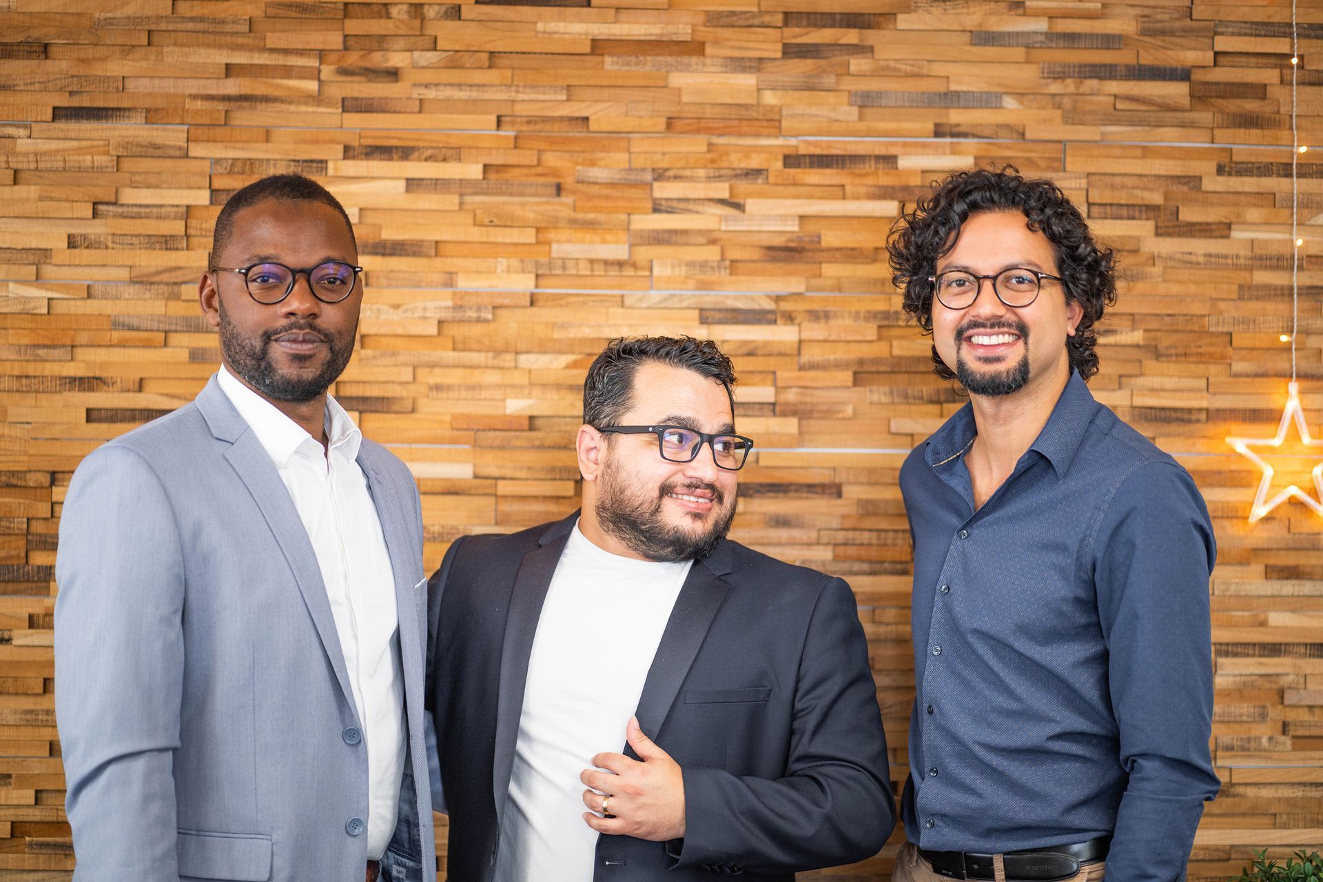 Trois hommes en chemise et veste posant derrière un mur brun