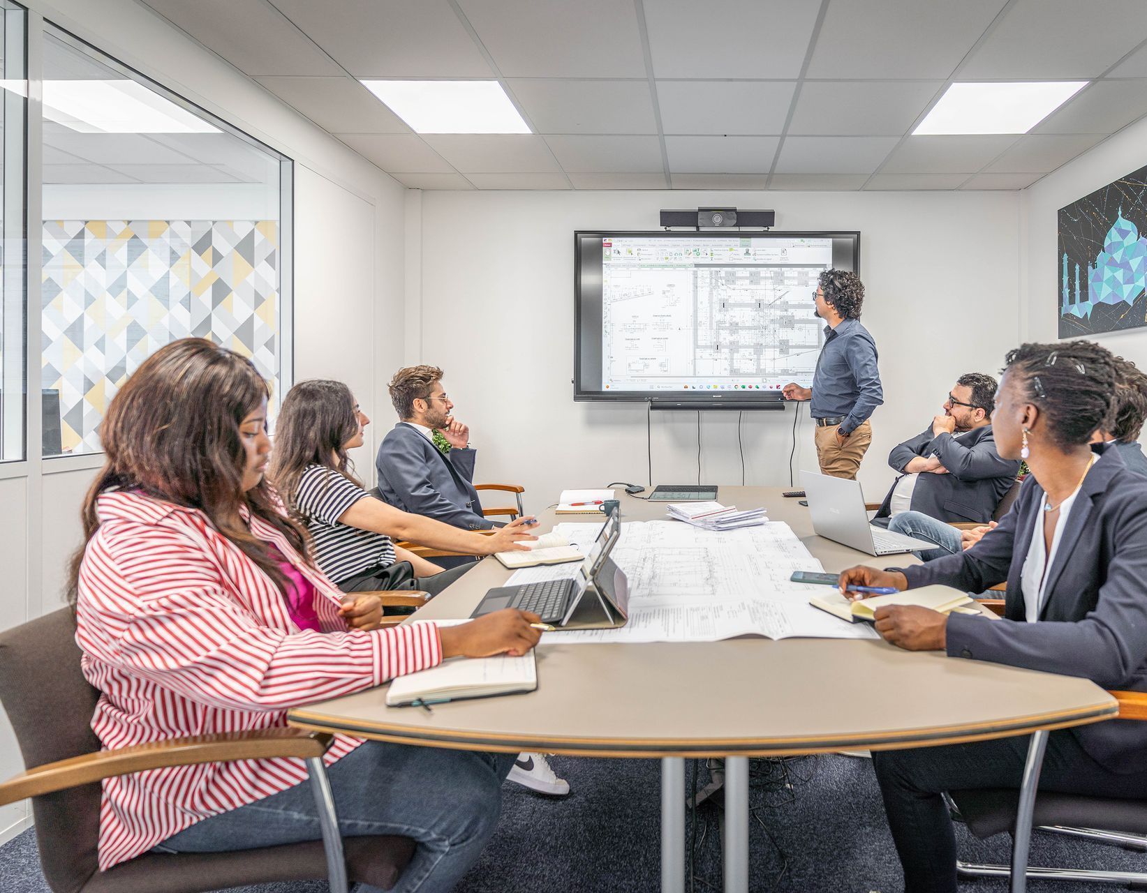 Équipe de bexconsult en réunion autour d'une table où l'un d'entre eux présente des plans sur un écran fixé au mur
