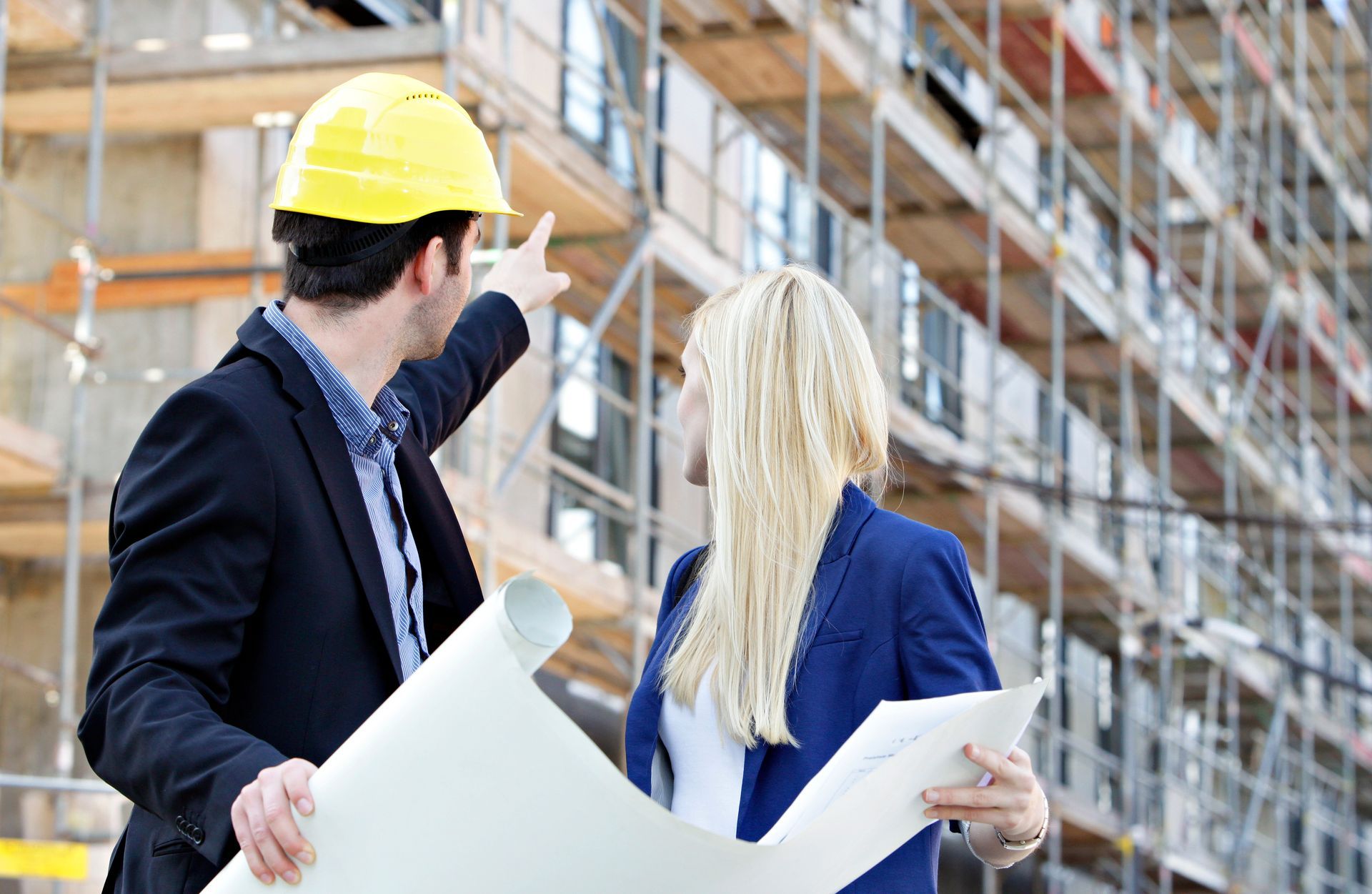 Un homme et une femme tournés de dos tenant des plans de construction devant un immeuble en chantier