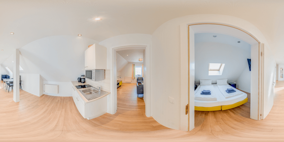 360-Grad-Ansicht einer lichtdurchfluteten Wohnung. Küchenbereich links, Schlafzimmer mit Doppelbett durch eine offene Tür rechts.
