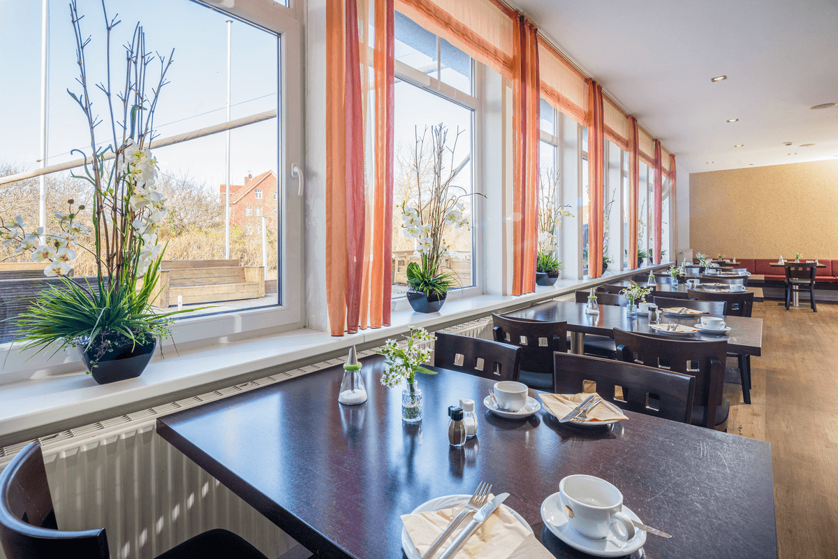 Esszimmer mit gedeckten Tischen, großen Fenstern und orangefarbenen Vorhängen.