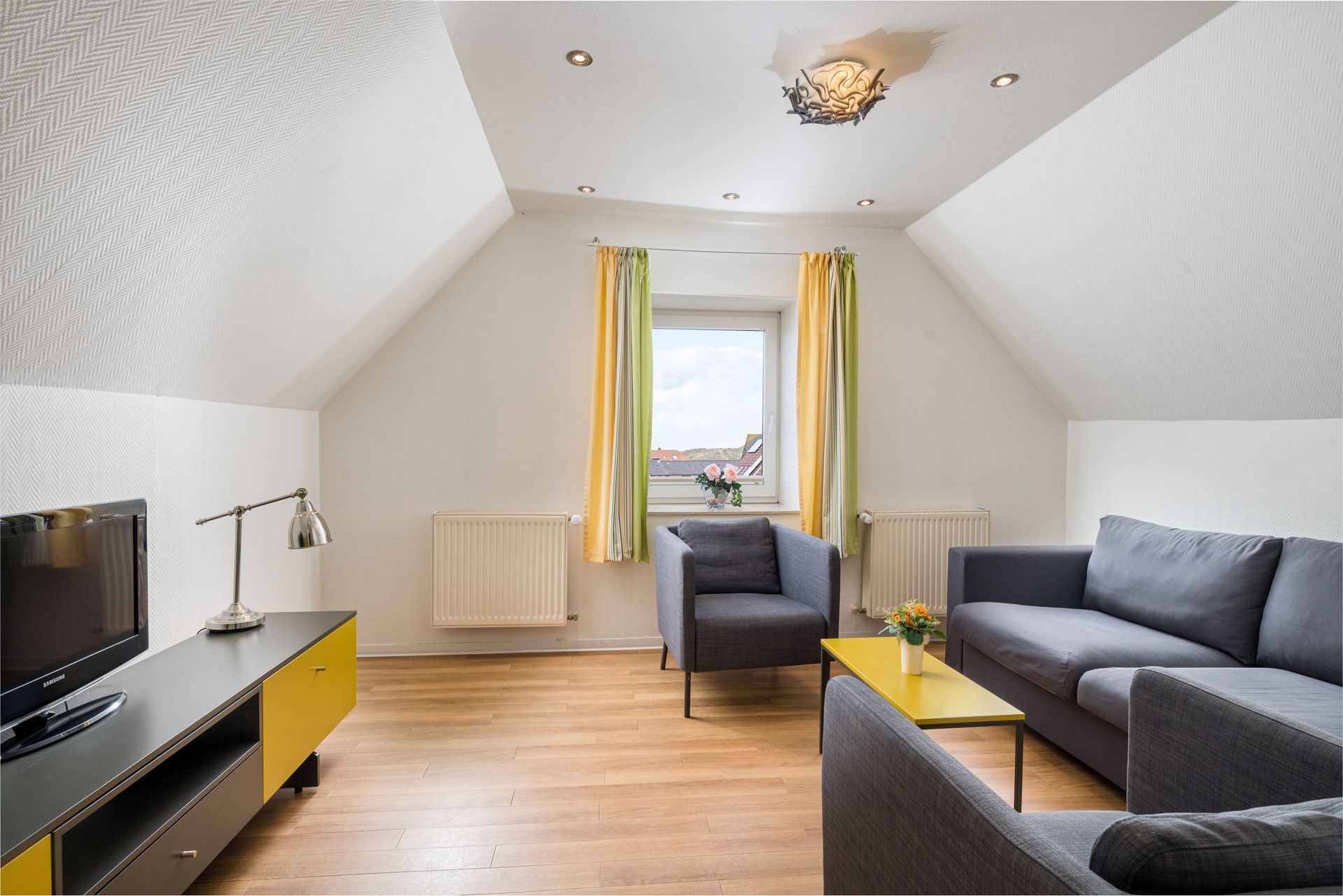 Wohnzimmer mit grauem Sofa, gelben Akzenten und Fenster mit farbenfrohen Vorhängen.