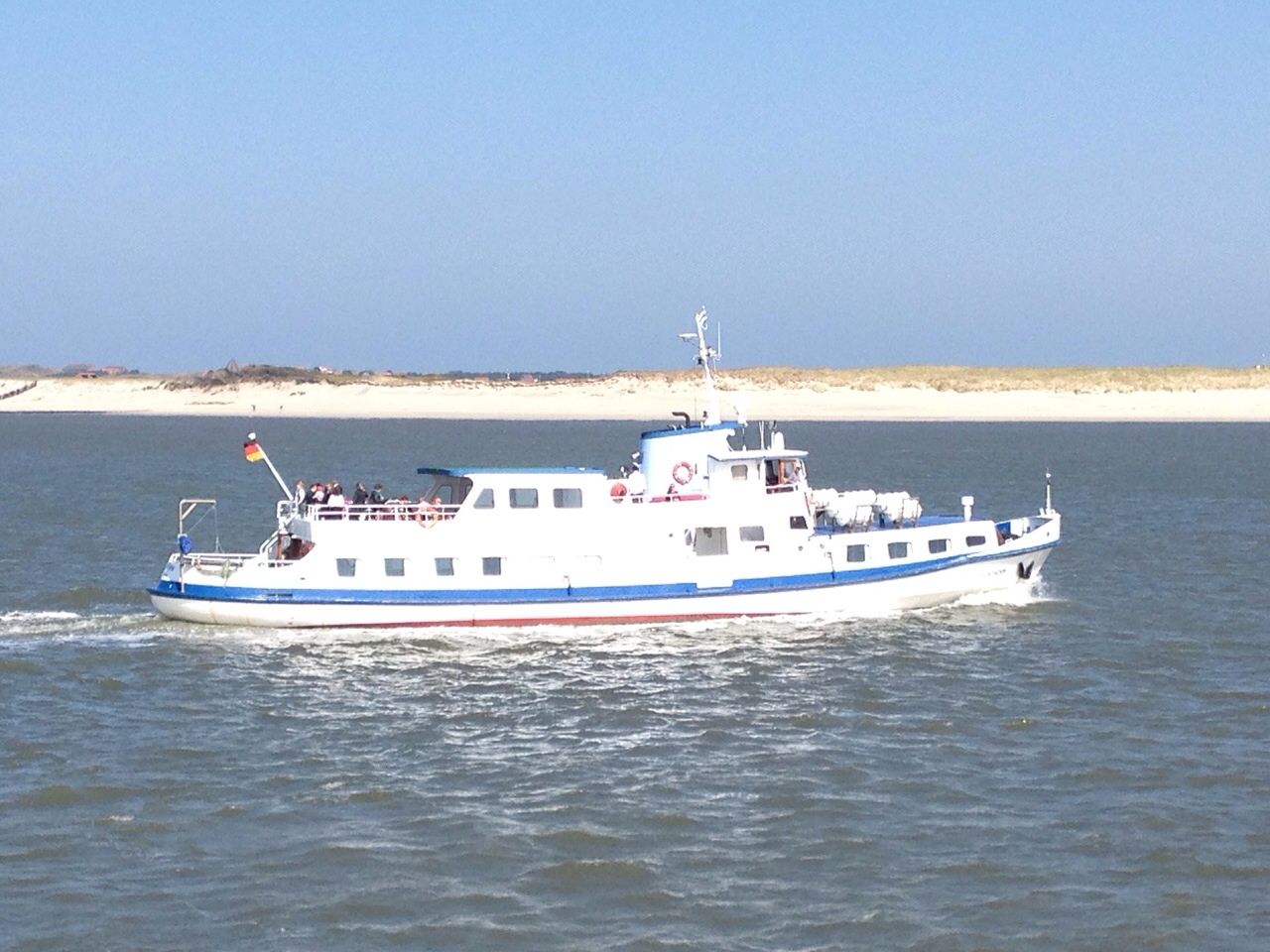 Weißes Passagierboot auf blauem Wasser, deutsche Flagge, in der Nähe eines Sandstrandes.