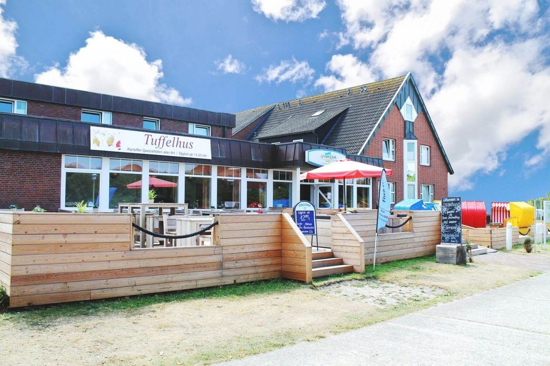 Restaurant mit Holzterrasse und rotem Sonnenschirm unter blauem Himmel.