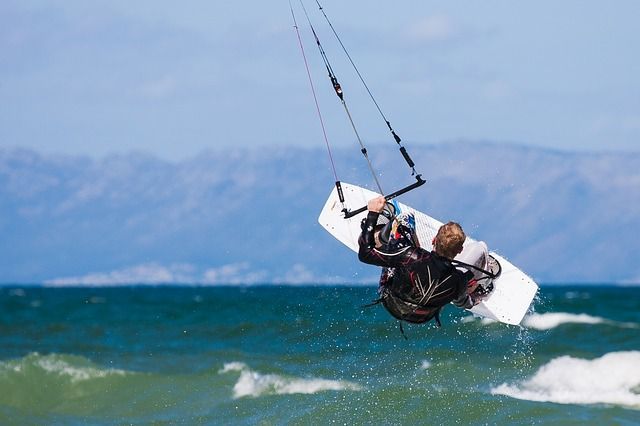 Ein Kitesurfer schwebt über dem Meer, hält sich an der Bar fest, der Kite über ihm. Blauer Himmel und Berge im Hintergrund.