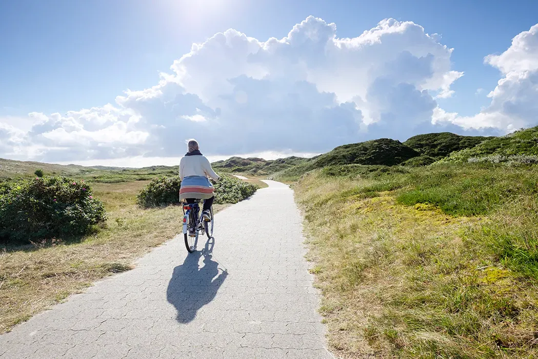 Eine Person fährt mit dem Fahrrad auf einem Pfad durch eine grasbewachsene Landschaft, strahlender Sonnenschein mit bauschigen Wolken.