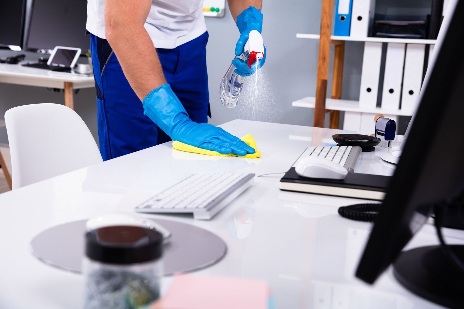 Eine Person mit blauen Handschuhen reinigt einen Schreibtisch mit einer Sprühflasche.