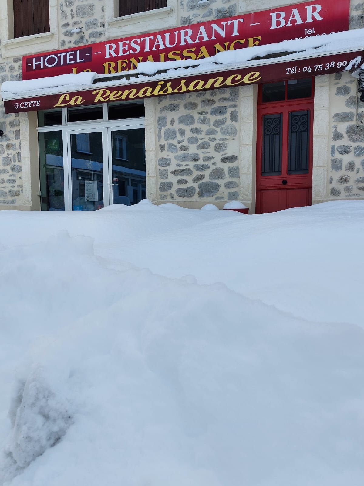 Entrée enneigée de l'hôtel, restaurant et bar La Renaissance ; porte rouge et enseigne visibles.