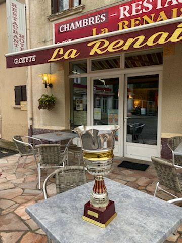 Le restaurant La Renaissance, avec un trophée sur une table en terrasse. Auvent marron, bâtiment beige et chaises en métal.