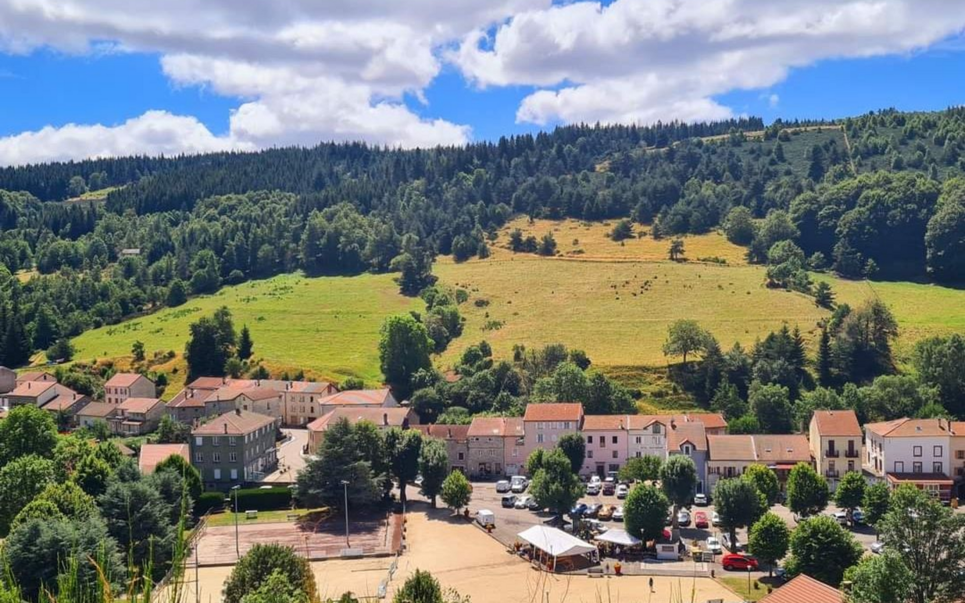 Village niché au pied d'une colline boisée, avec un champ verdoyant et un ciel bleu au-dessus.