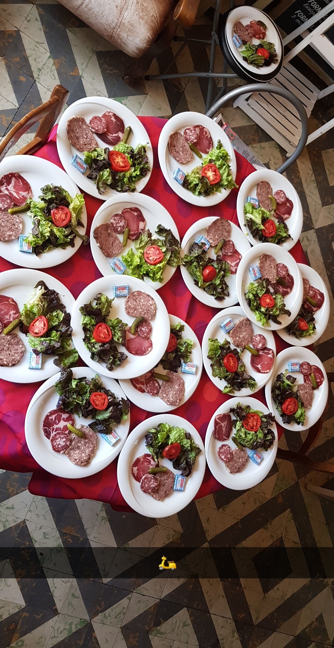 Des assiettes de viande, de salade et de tomates sont disposées sur une nappe rouge. Une table et une chaise sont visibles en arrière-plan.