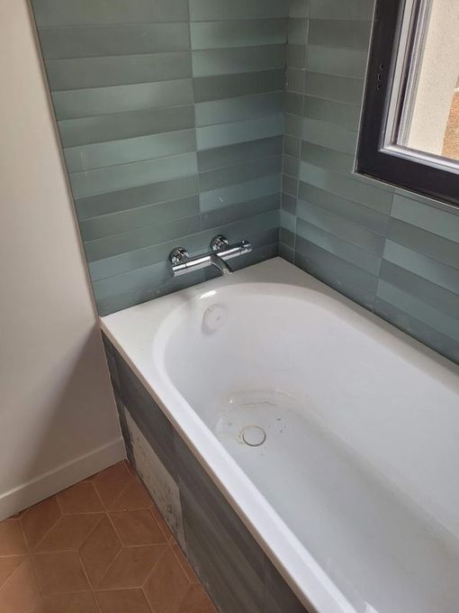 Petite salle de bains avec baignoire blanche, murs carrelés gris et fenêtre au-dessus de la baignoire.
