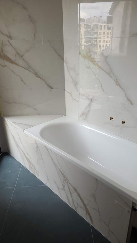 Salle de bains moderne avec baignoire blanche, murs carrelés imitation marbre, sol foncé et reflet de fenêtre.