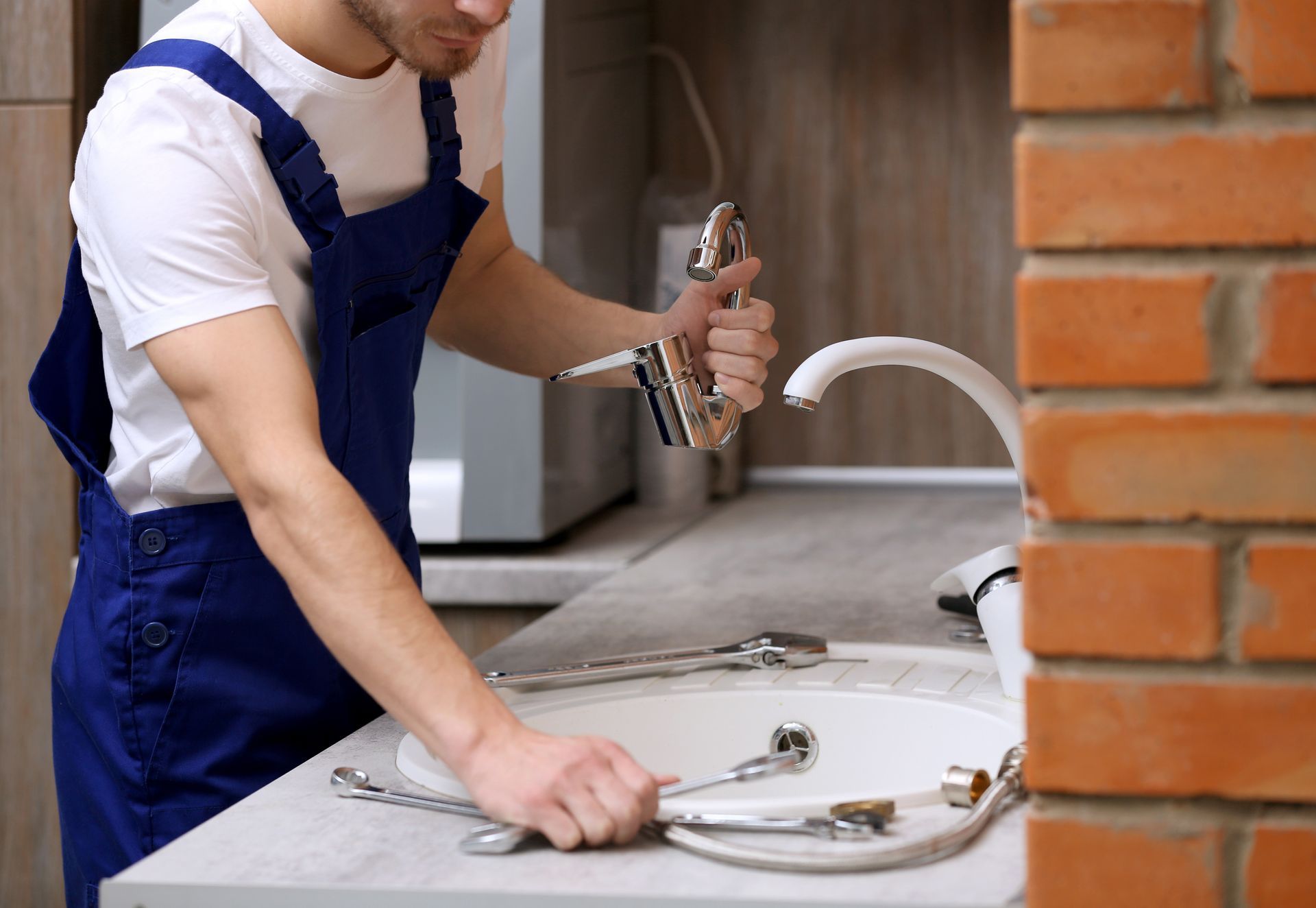 Un plombier en salopette bleue installe un robinet de cuisine à côté d'un mur de briques et d'un évier.