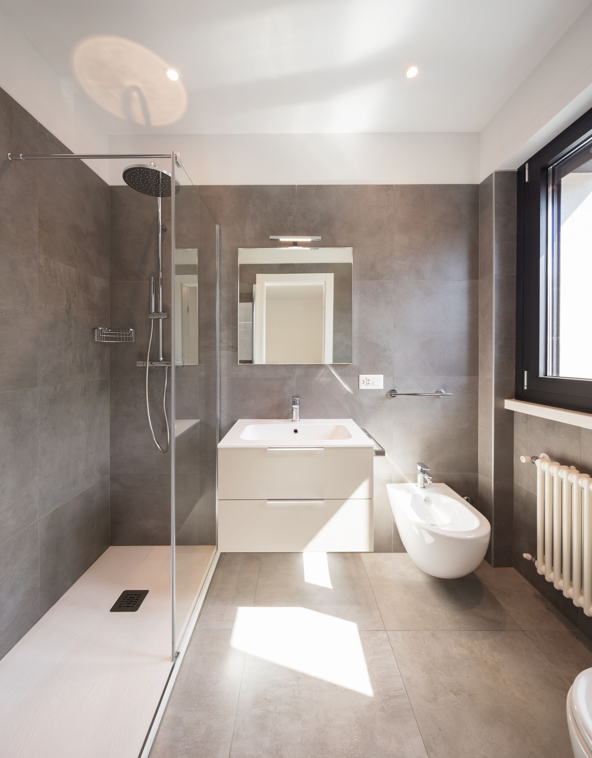Salle de bains moderne avec douche à l'italienne, meuble sous lavabo, toilettes et murs carrelés gris ; lumière naturelle abondante.