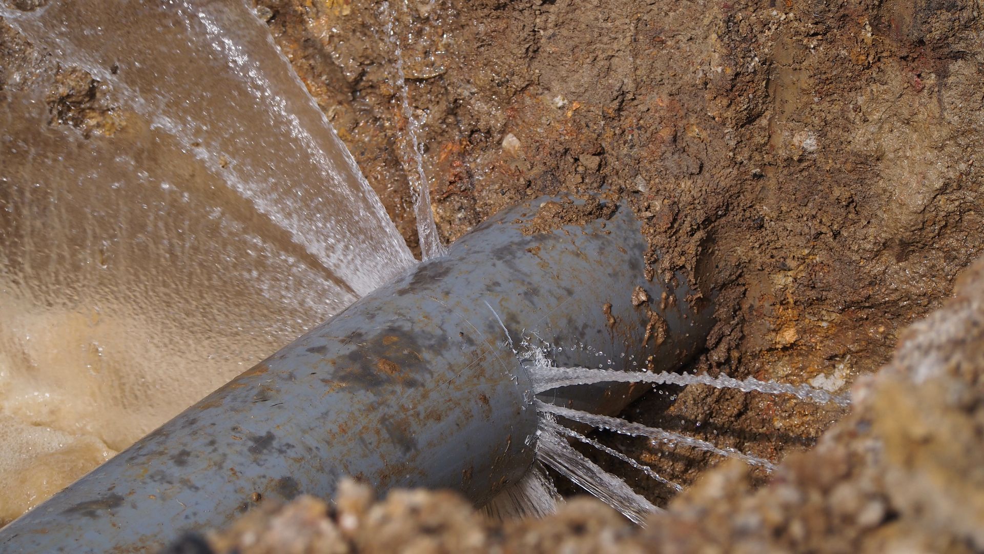 De l'eau jaillit d'un tuyau gris cassé dans une tranchée.