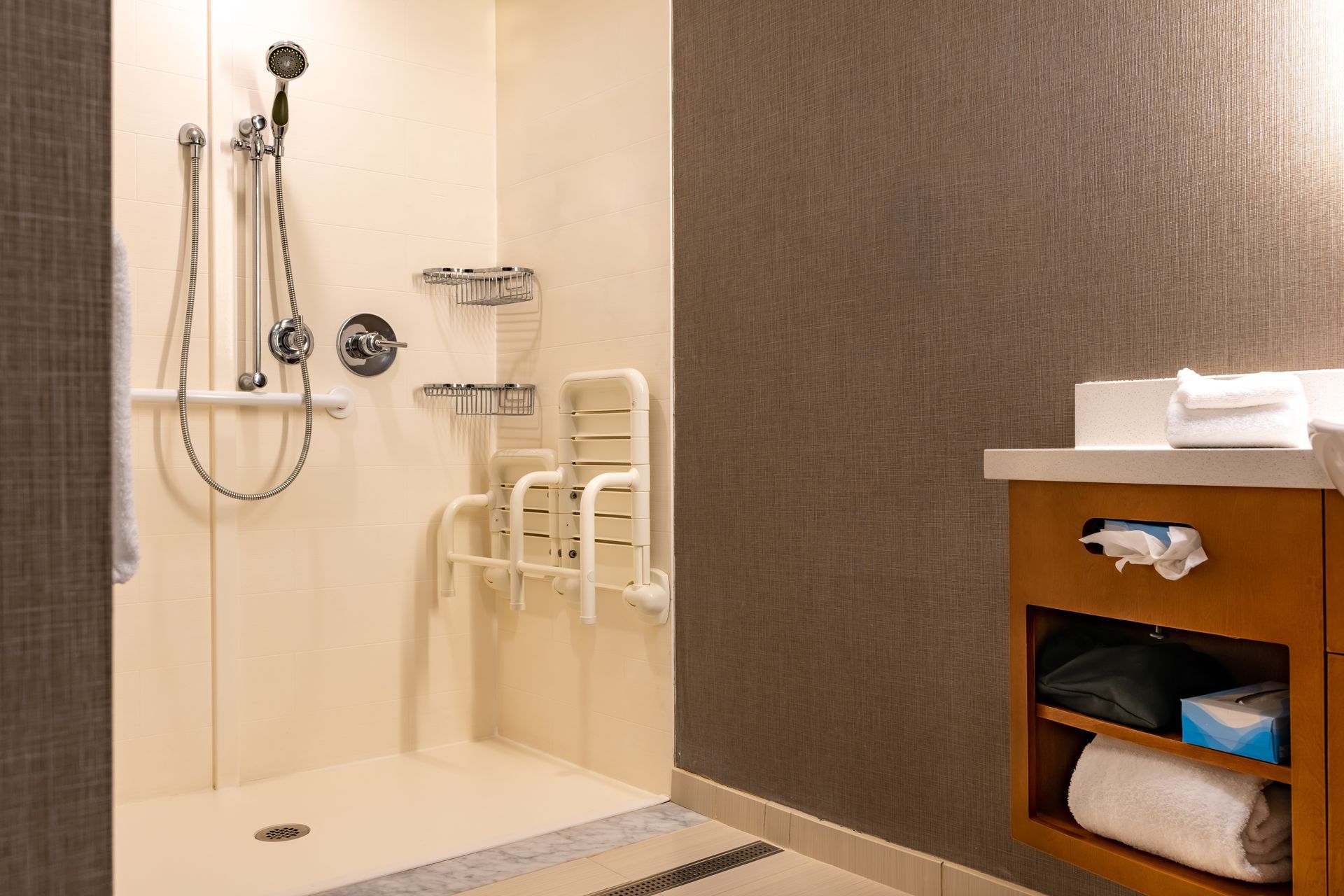 Salle de bains accessible avec douche, chaise, serviettes et meuble sous lavabo.