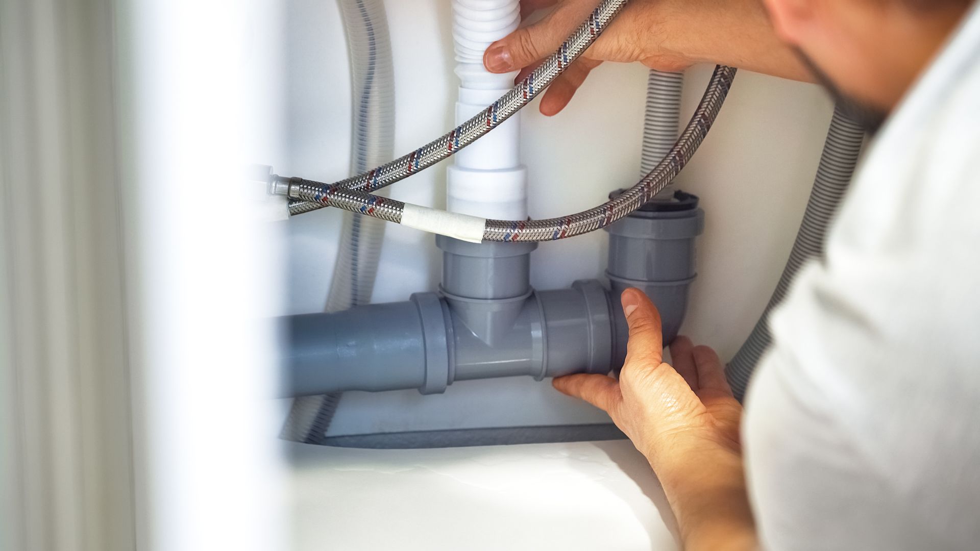 Des mains inspectent les tuyaux de plomberie gris sous un évier.